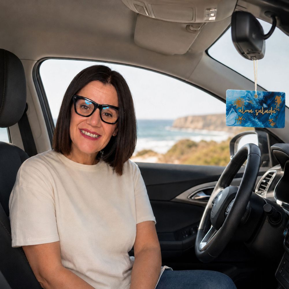 Mari Carmen, CEO y experta en aromas de D'Arome, en el asiento del conductor de su coche con el ambientador Alma Salada de LoeS colgando del retrovisor frente a la playa – D'Arome