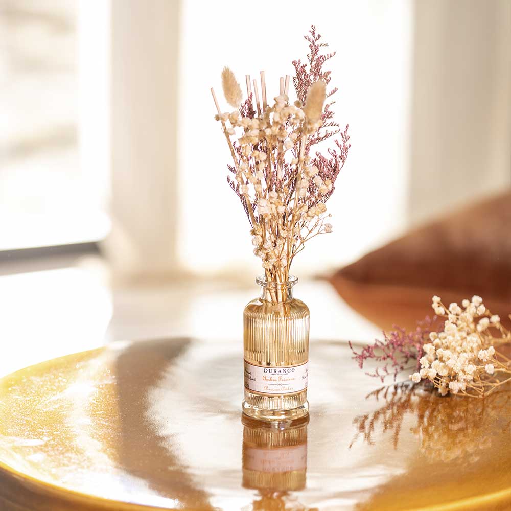 Difusor de aromas Ámbar Precioso de Durance con ramo de flores secas iluminado por una luz cálida sobre una superficie dorada, junto a un ramillete suelto.