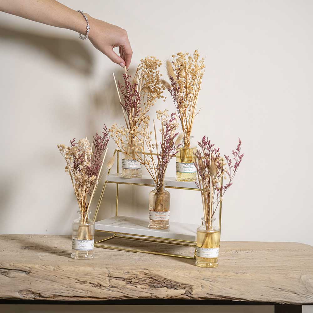 Colección de ambientadores mikado con flores secas naturales de la marca Durance sobre un elegante expositor de mármol y dorado. Mano poniendo varillas.