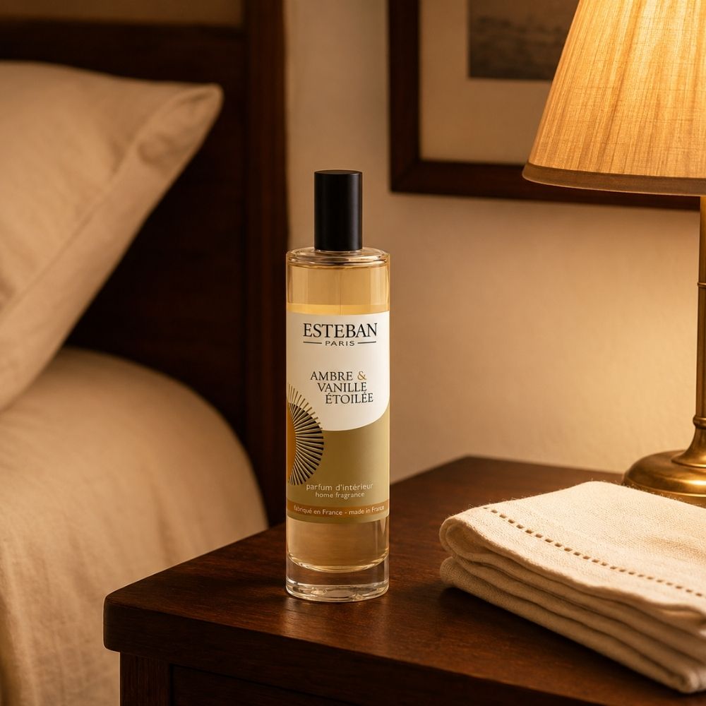 Ambientador spray Ambre et Vanille Étoilée Esteban París sobre mesilla de dormitorio junto a lámpara y toallas – D'Arome