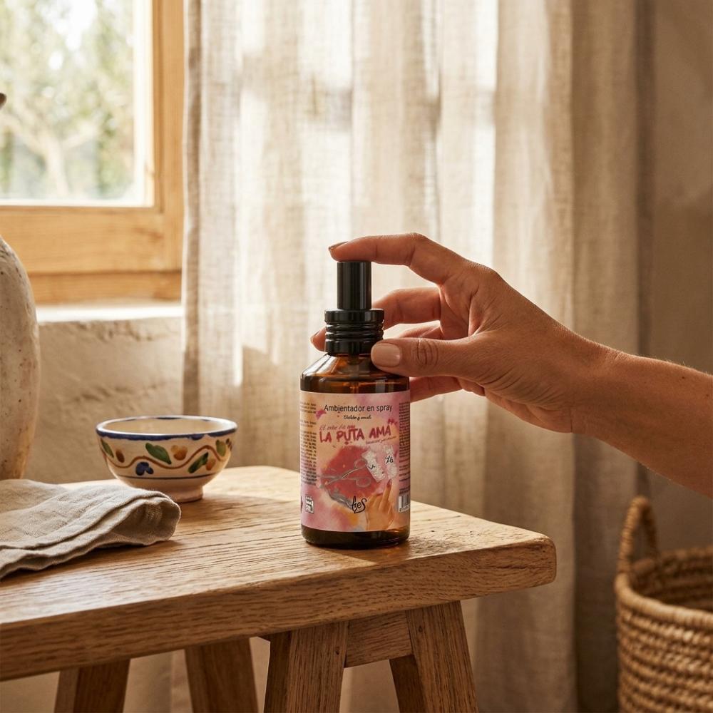 Mano cogiendo el spray ambientador La Puta Ama LoeS violeta y musk en taburete de madera con luz natural, ambientador hogar aroma violeta – D'Arome