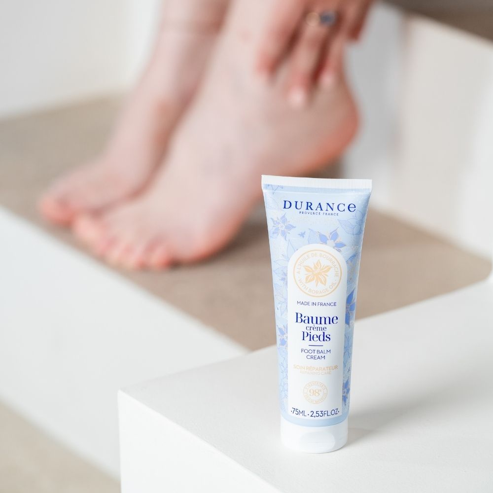 Bálsamo crema de pies con aceite de borraja Durance sobre escalón blanco minimalista con pies desenfocados al fondo – D'Arome