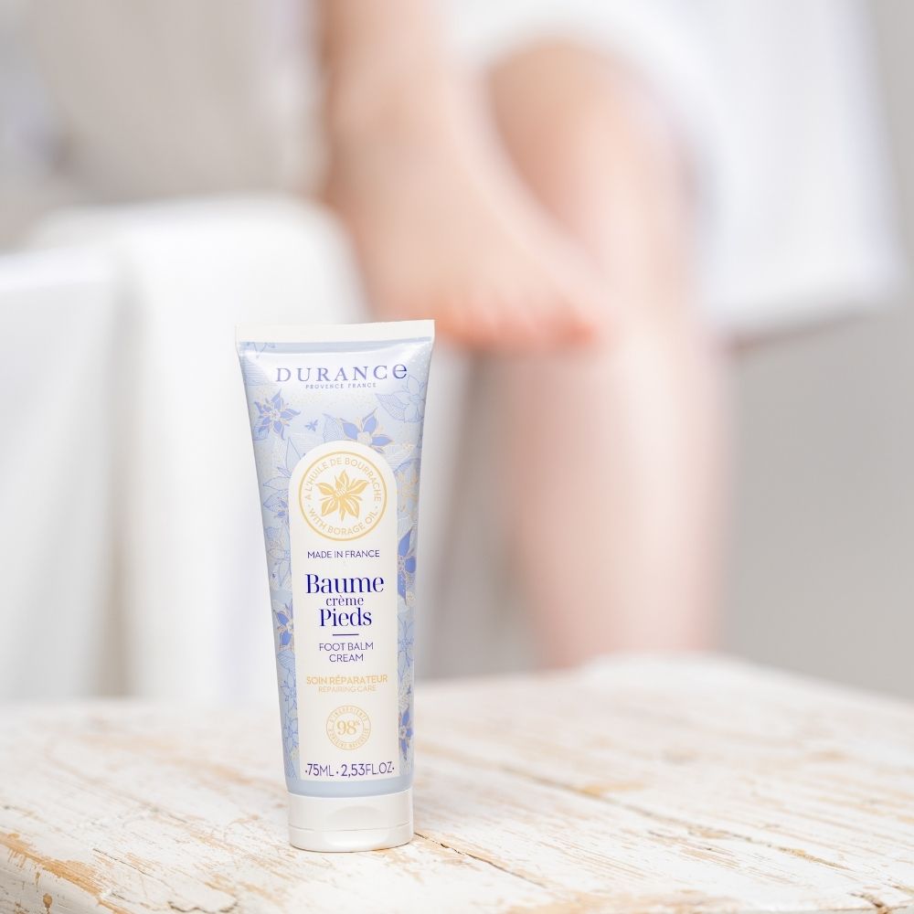 Bálsamo crema de pies con aceite de borraja Durance sobre mesa de madera envejecida con piernas desenfocadas de fondo – D'Arome