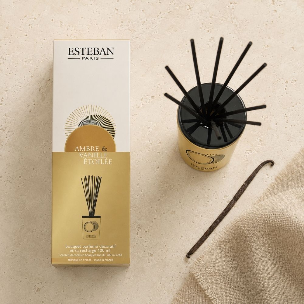 Vista cenital del bouquet perfumado Ambre & Vanille Étoilée de Esteban Paris junto a su caja, una vaina de vainilla y un textil de lino sobre piedra clara – D'Arome