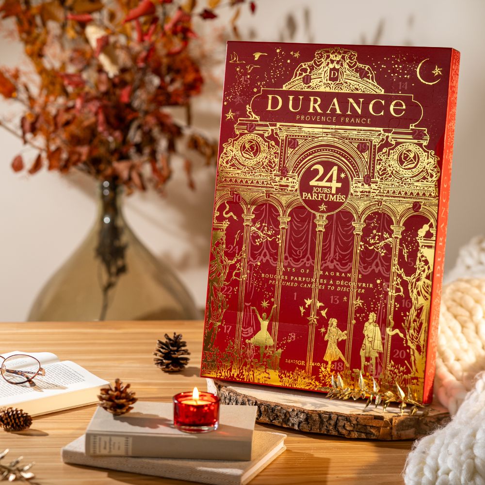 Calendario de adviento Durance 2026 con 24 velas perfumadas apoyado en mesa de madera junto a libros, vela roja encendida y piñas, ambientación otoñal – D'Arome