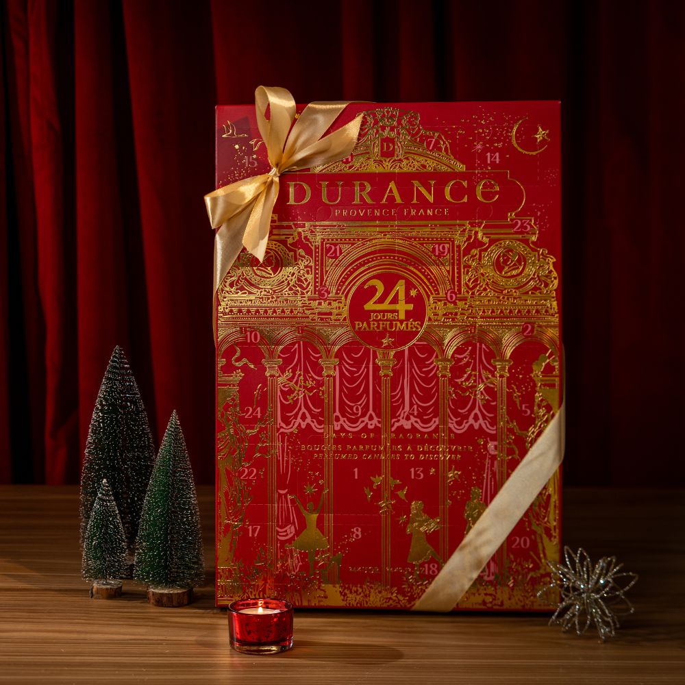 Calendario de adviento Durance 2026 cerrado, caja roja con 24 velas perfumadas y lazo de raso dorado, sobre fondo de telón de un teatro donde ver ópera rojo ambiente navideño con arbolitos decorativos. Se ve el portavelas rojo con vela de te encendida – D'Arome