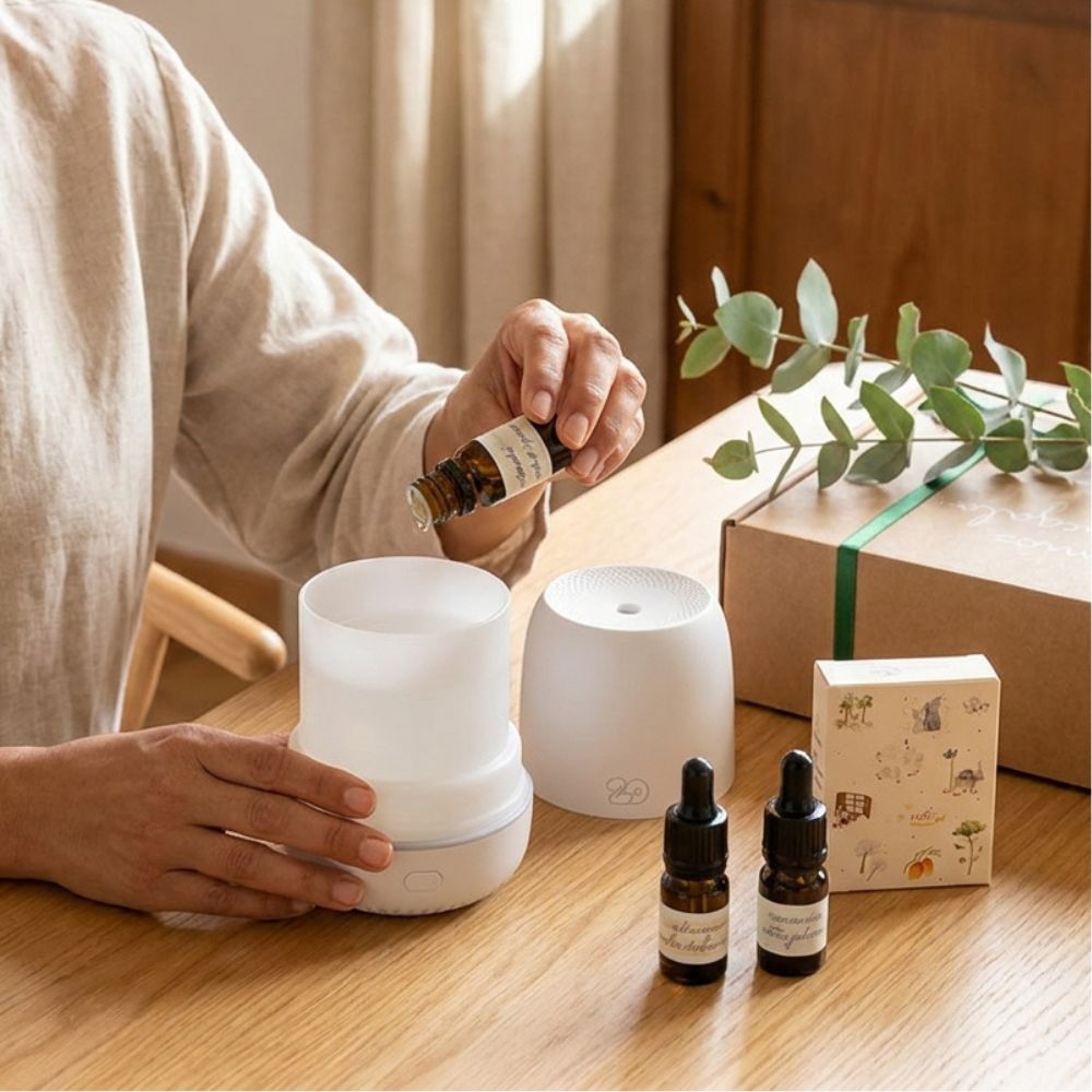 Uso del cofre regalo original de D'Arome: echando gotas de mini esencia LoeS en el difusor de aromas Sira blanco junto a caja kraft regalo con lazo verde en salón – D'Arome