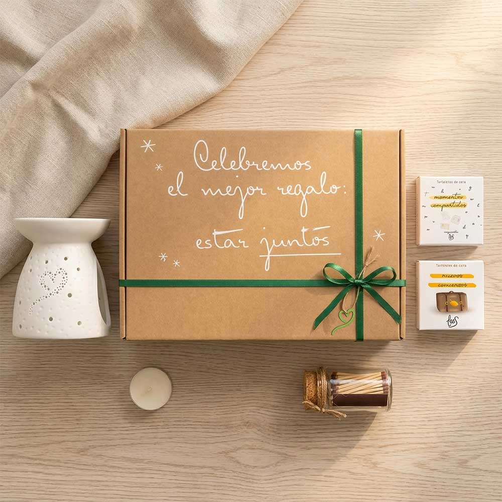 Vista cenital del cofre regalo para profesora de D'Arome con caja kraft cerrada con lazo verde, quemador corazón de cerámica, tartaletas de cera Momentos Compartidos y Nuevos Comienzos de LoeS y cerillas decorativas sobre tela de lino con eucalipto – D'Arome