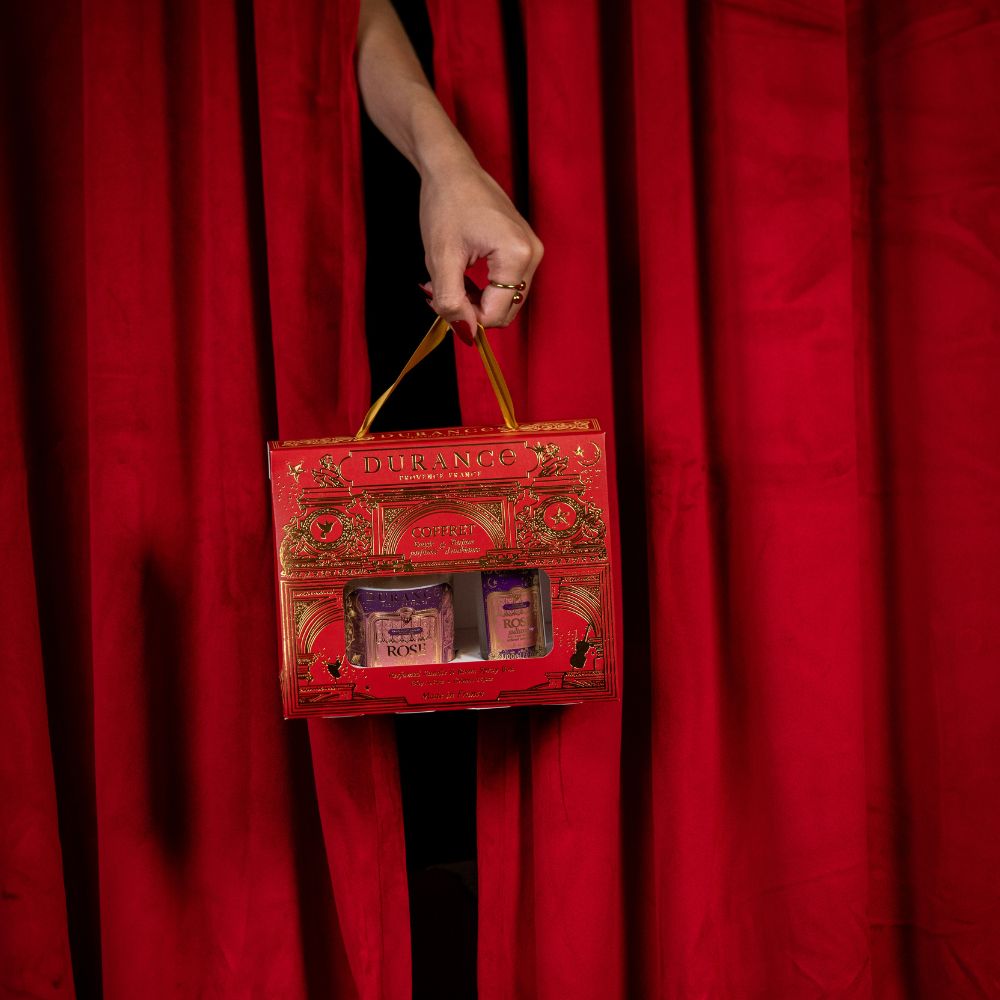 Mano con manicura roja y anillo dorado sujetando el cofre regalo vela y spray Rosa Sultana de Durance por su asa dorada, fondo de cortina roja de teatro – D'Arome