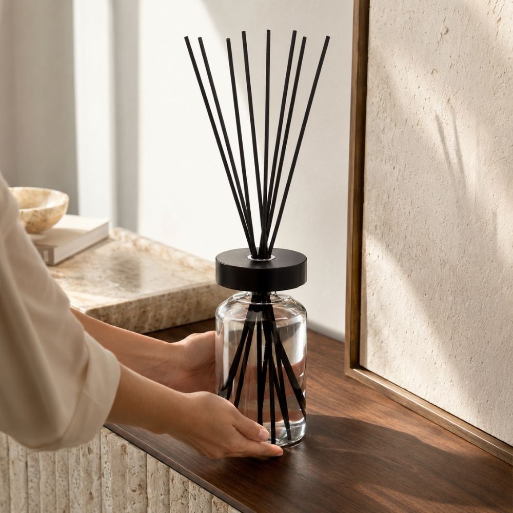 Manos colocando el difusor mikado Figue Noire de Esteban Paris sobre una repisa de madera junto a una pared de piedra travertino – D'Arome