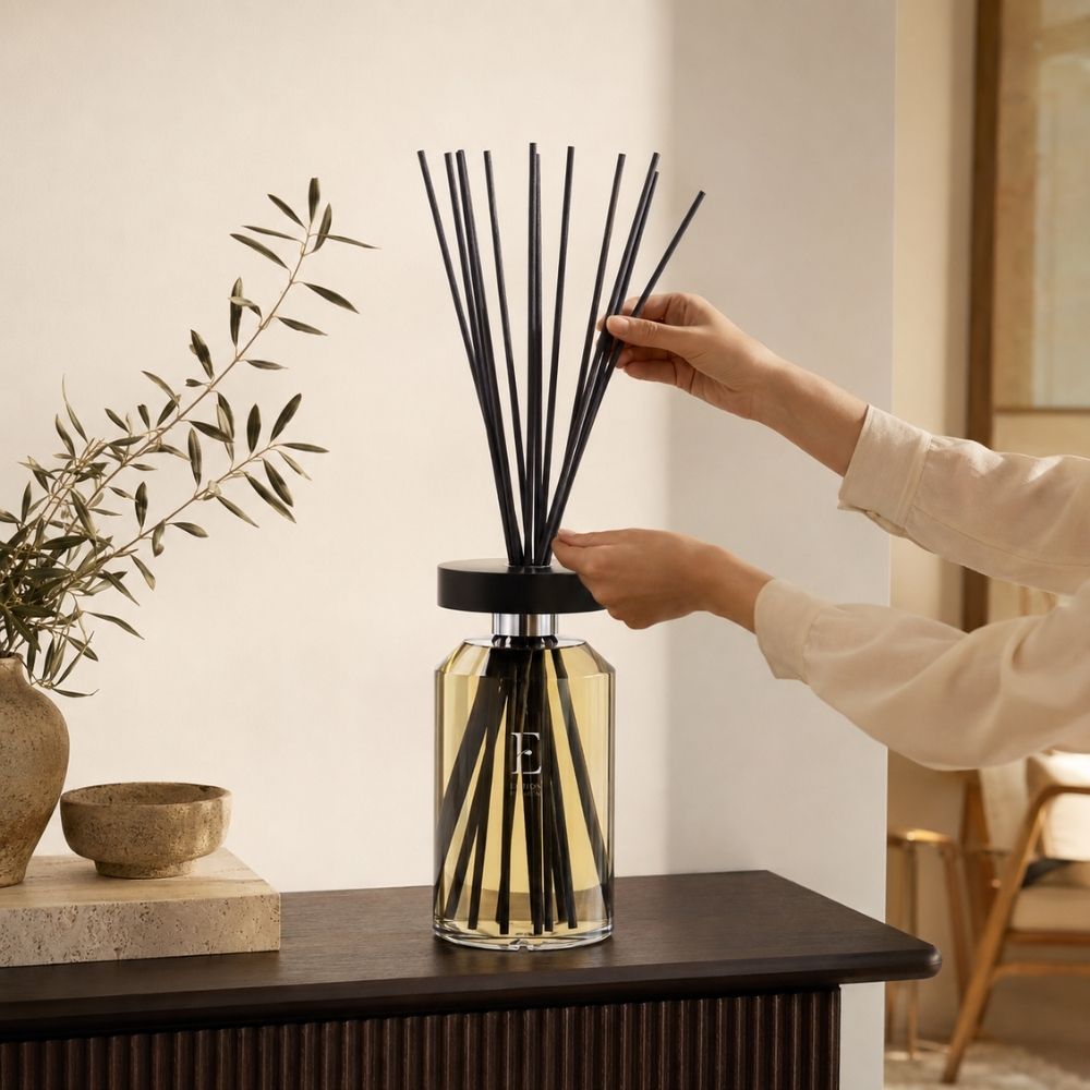 Manos ajustando las varillas del difusor mikado Néroli de Esteban Paris sobre un mueble oscuro, junto a una rama de olivo y un cuenco de piedra – D'Arome