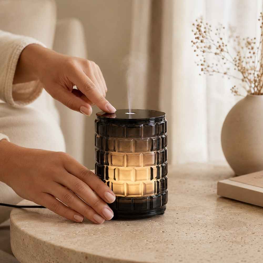Detalle de manos activando el difusor eléctrico celebration de cristal ahumado Maison Berger con vapor aromático visible – D'Arome