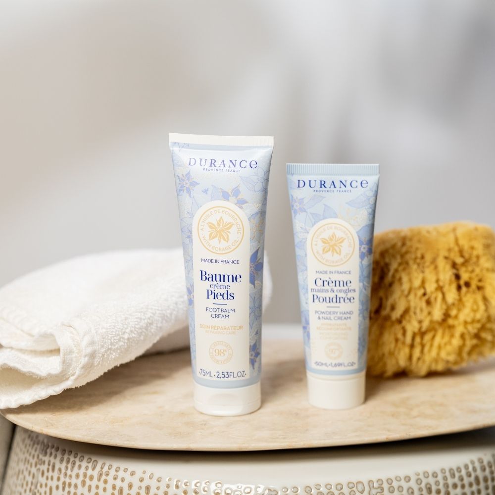 Dúo de bálsamo crema de pies y crema de manos y uñas Poudrée con aceite de borraja Durance sobre repisa junto a toalla y esponja – D'Arome