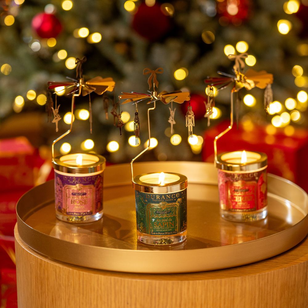 Tres velas carrusel giratorias Durance encendidas sobre mesa dorada con árbol de Navidad de fondo: Rosa Sultana, Al Pie del Abeto y Cereza Aterciopelada, 180gr cada una – D'Arome