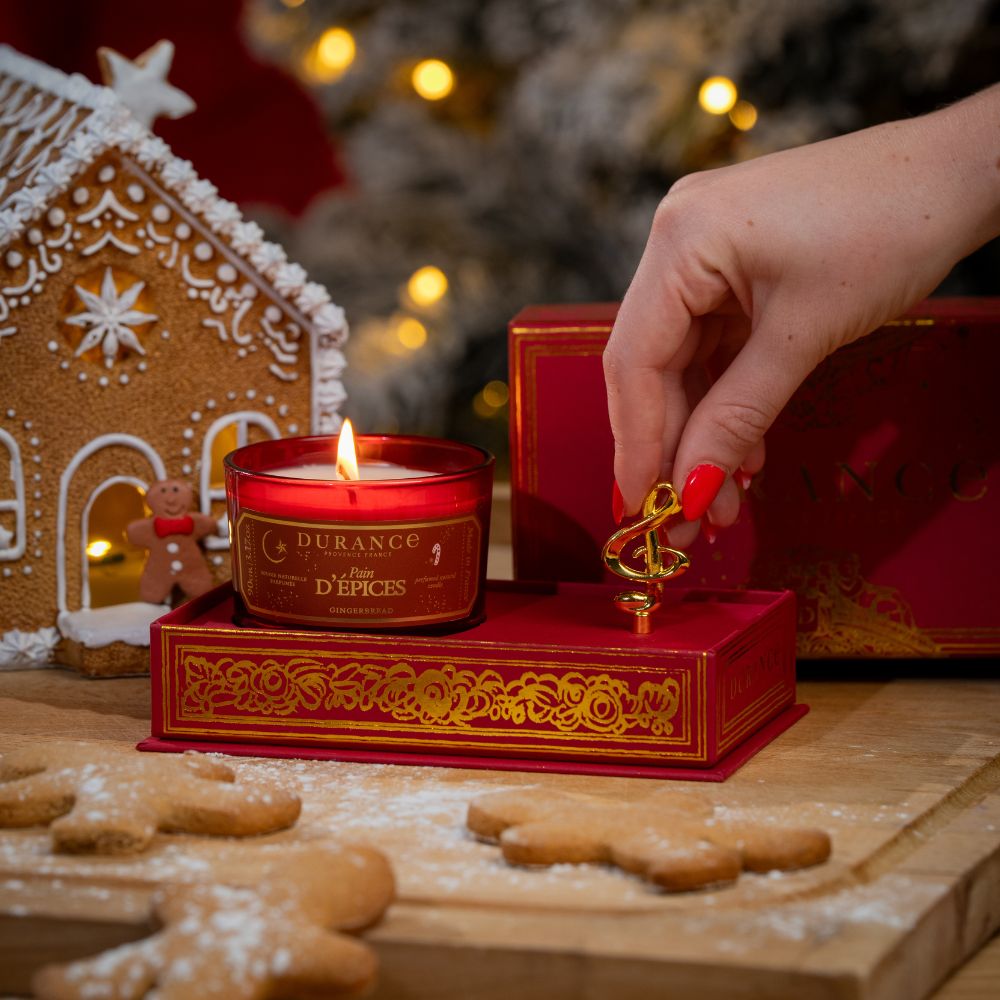 Mano girando la clave de sol dorada de la caja de música de Durance con vela Pan de Especias 90gr encendida, junto a casita de jengibre y galletas navideñas – D'Arome