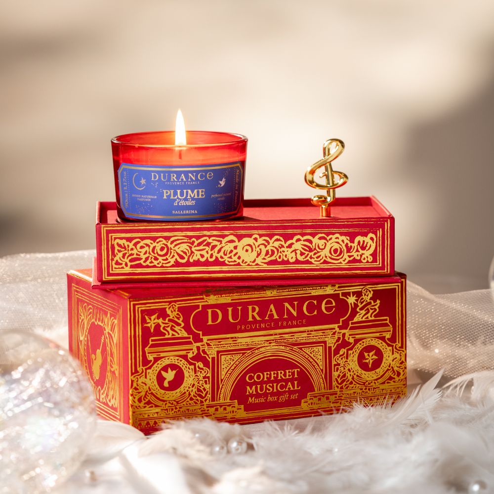 Vela Pluma de Estrellas 90gr de Durance encendida sobre la caja de música Coffret Musical apilada, con clave de sol dorada y ambientación de plumas blancas estilo bailarina – D'Arome