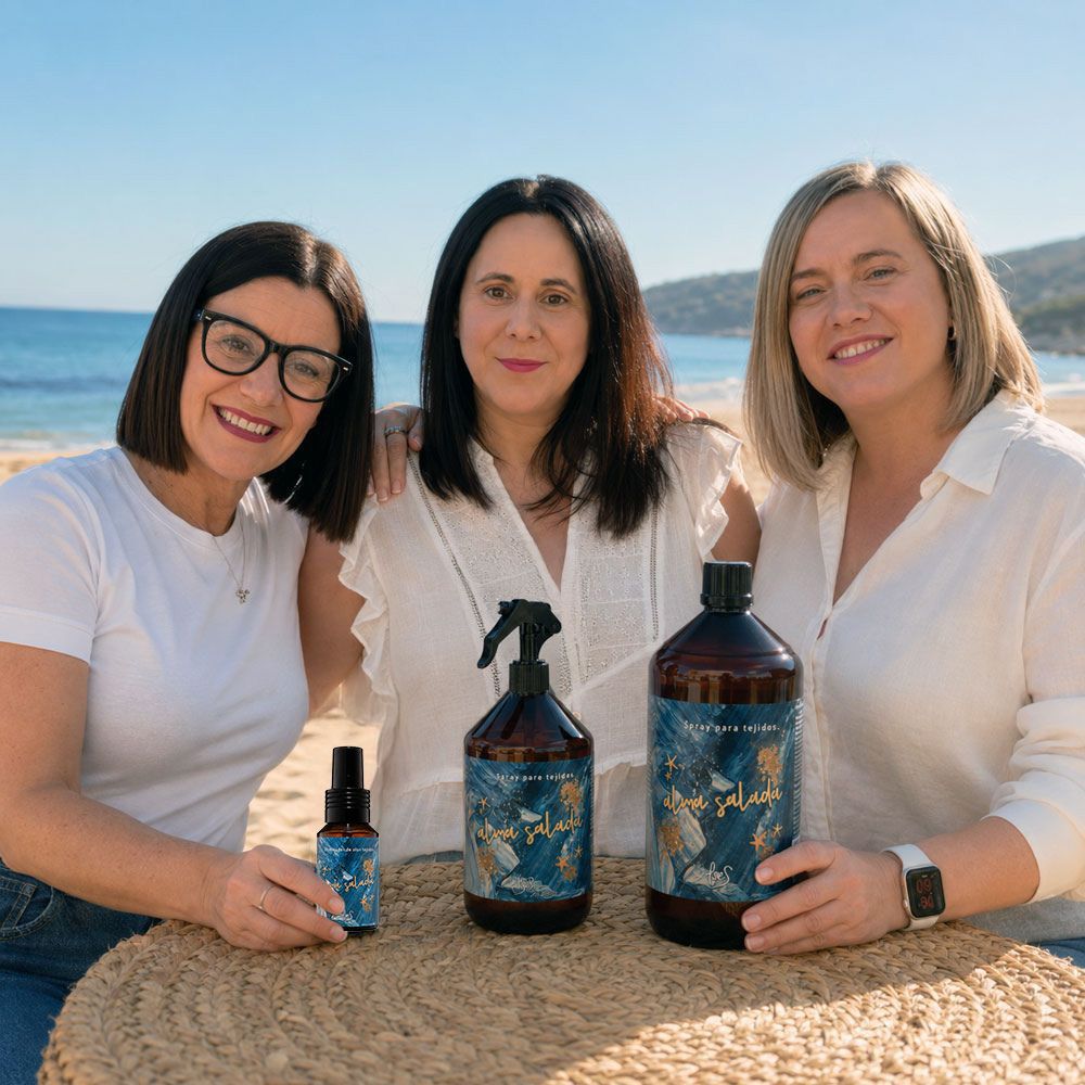 Las tres hermanas Darome fundadoras de D'Arome —Mari Carmen (CEO y experta en aromas), Paqui y Ana Belén (cofundadoras)— sostienen los tres formatos del spray textil Alma Salada de LoeS (mini 50 ml, 500 ml y recambio de 1 litro) en una playa mediterránea – D'Arome