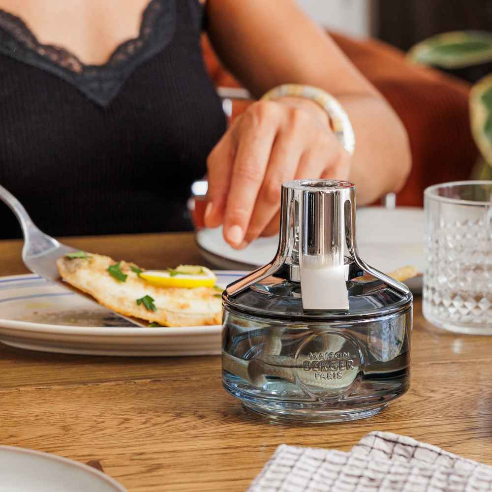 Lámpara catalítica Identity Maison Berger en cristal azul gris sobre mesa de cocina junto a un plato de pescado, ideal para eliminar el mal olor despues de cocinarlo – D'Arome