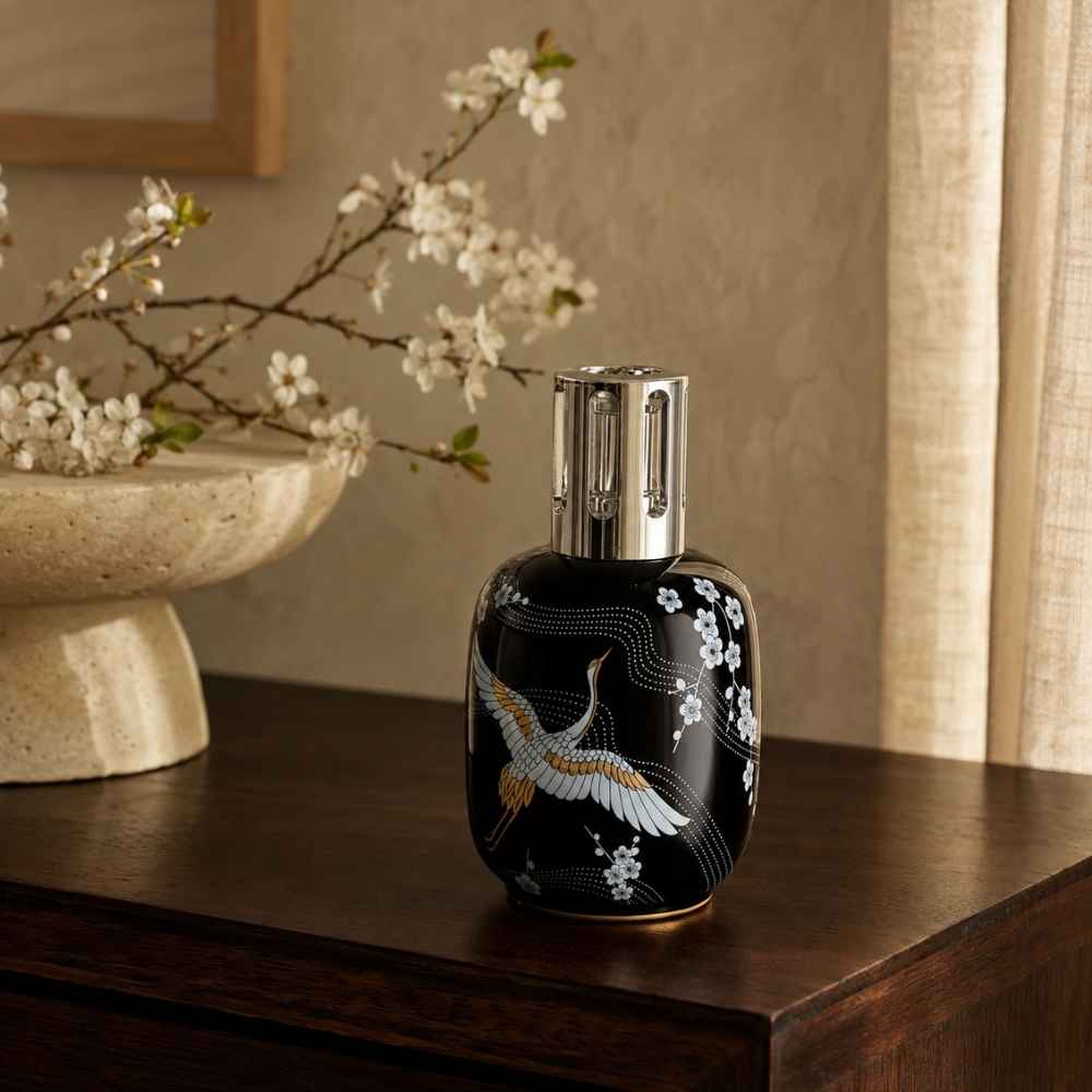 Lámpara catalítica Sakura Negra Maison Berger con grulla, embellecedor plateado y flores blancas sobre cómoda de madera oscura – D'Arome