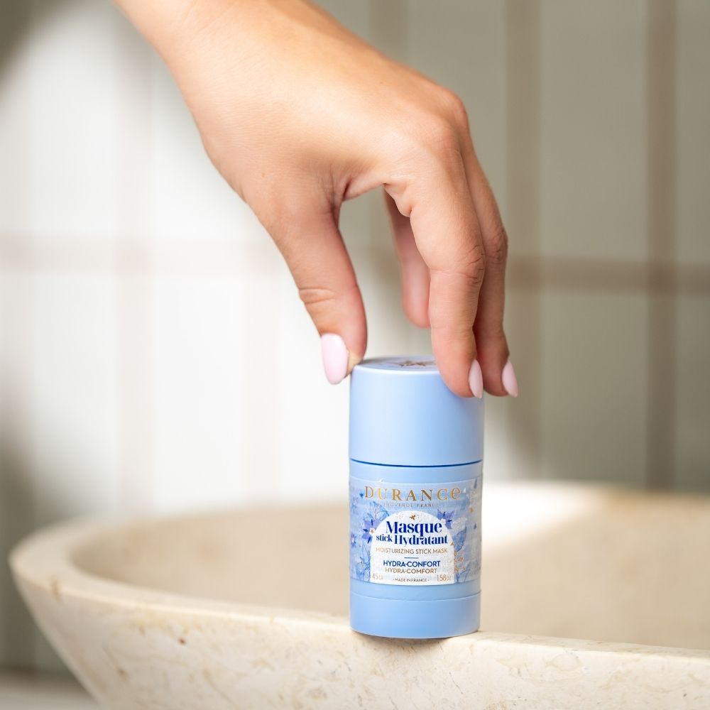Mano cogiendo la mascarilla hidratante en stick con aceite de borraja Durance desde el borde de un lavabo de piedra – D'Arome