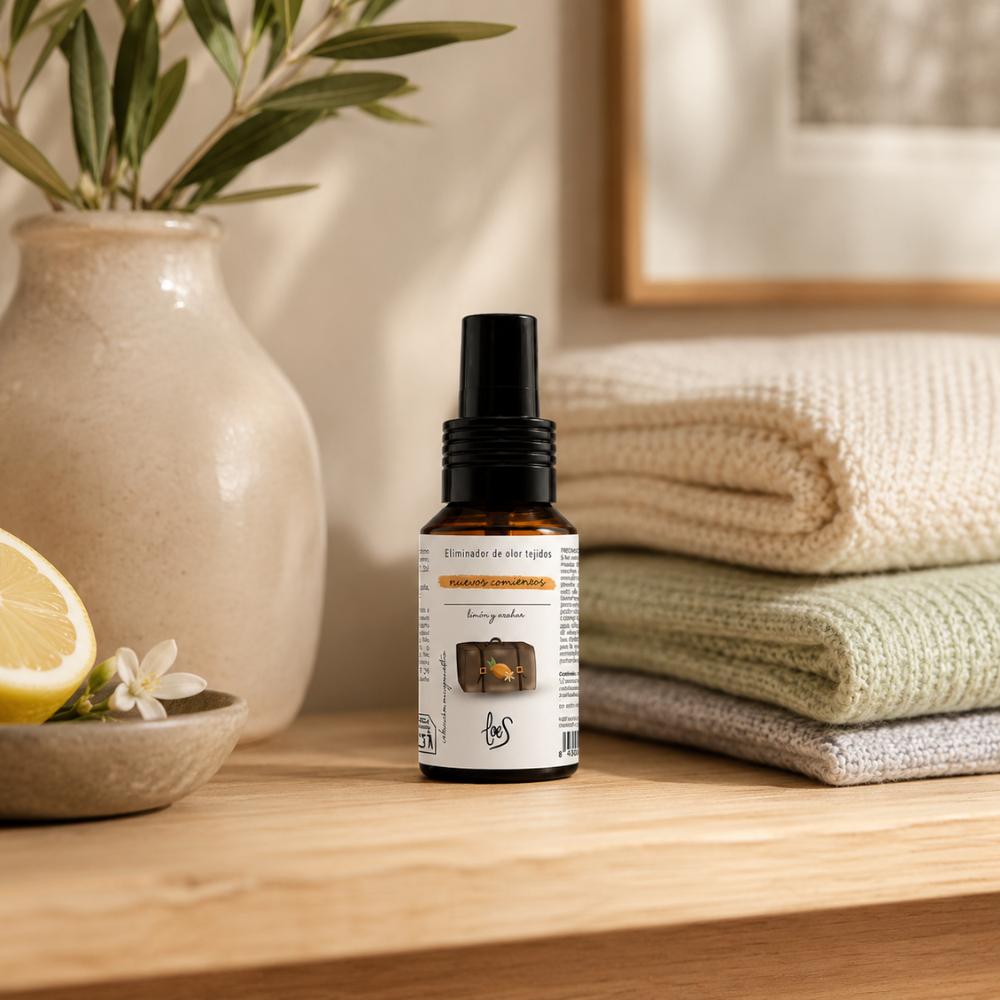 Mini spray textil eliminador de olores Nuevos Comienzos 50ml de LoeS con aroma a limón y azahar junto a limón cortado y flor de azahar sobre estante con toallas dobladas – D'Arome