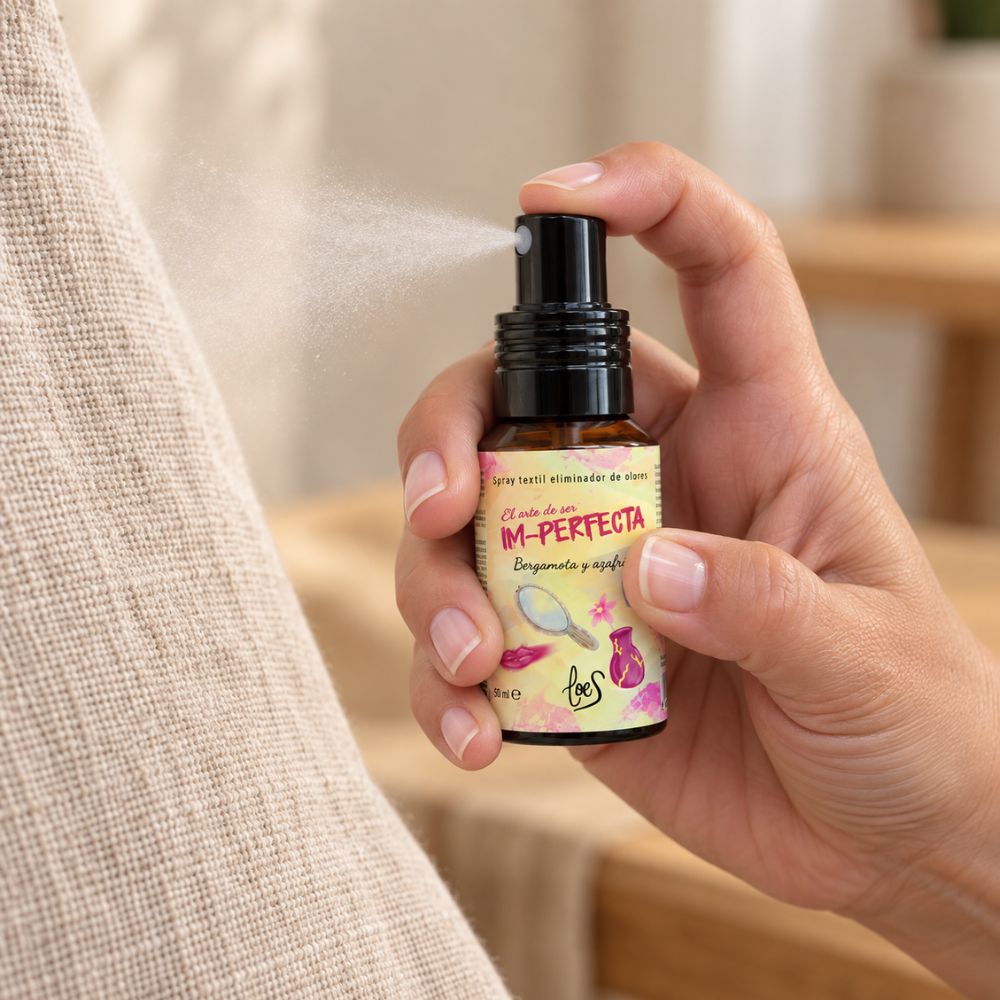 Detalle de una mano pulverizando el mini spray textil Im-Perfecta de LoeS sobre un cojín de lino, eliminando olores al instante – D'Arome