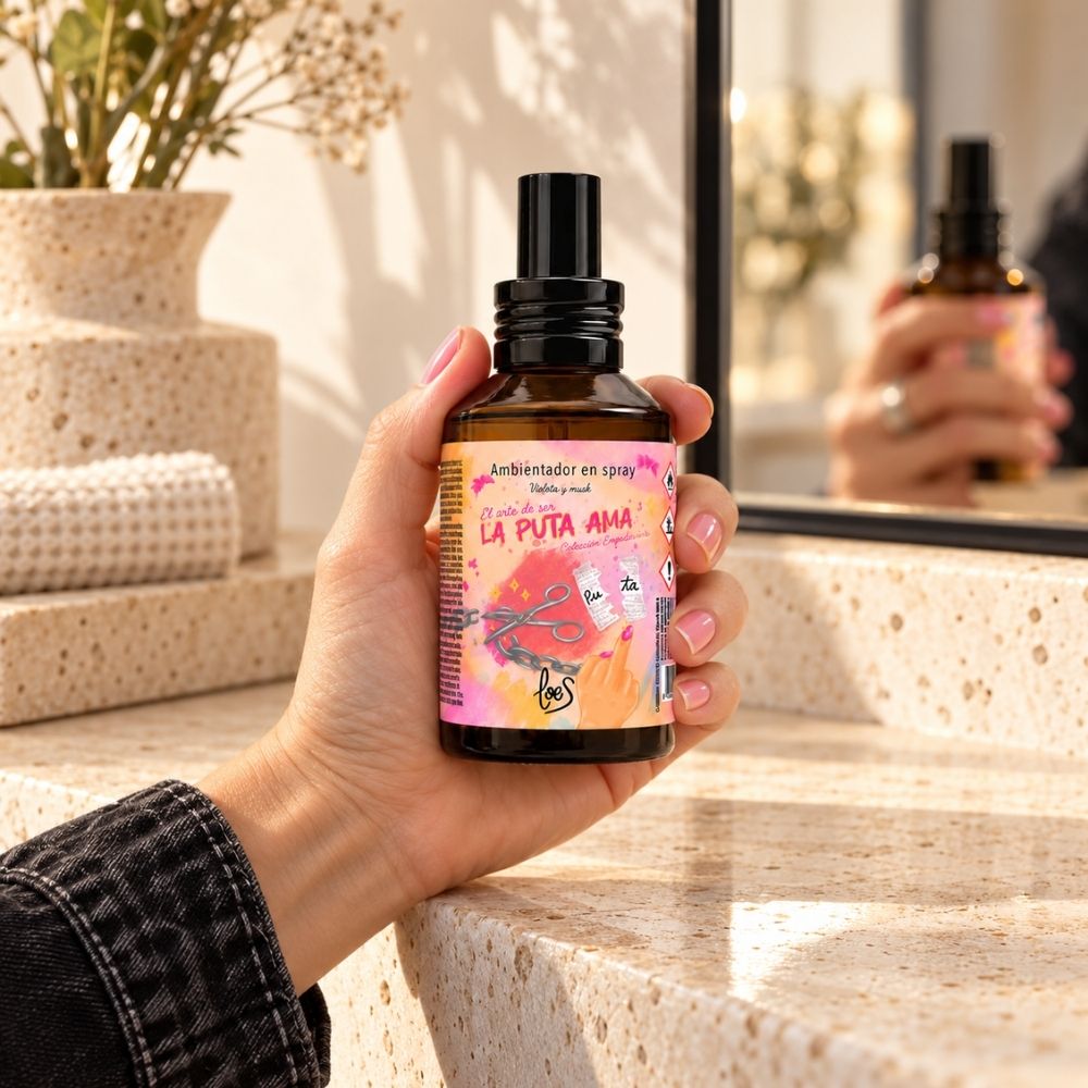 Mano sujetando el spray ambientador para el hogar La Puta Ama de LoeS junto al espejo, aroma a violeta y musk – D’Arome