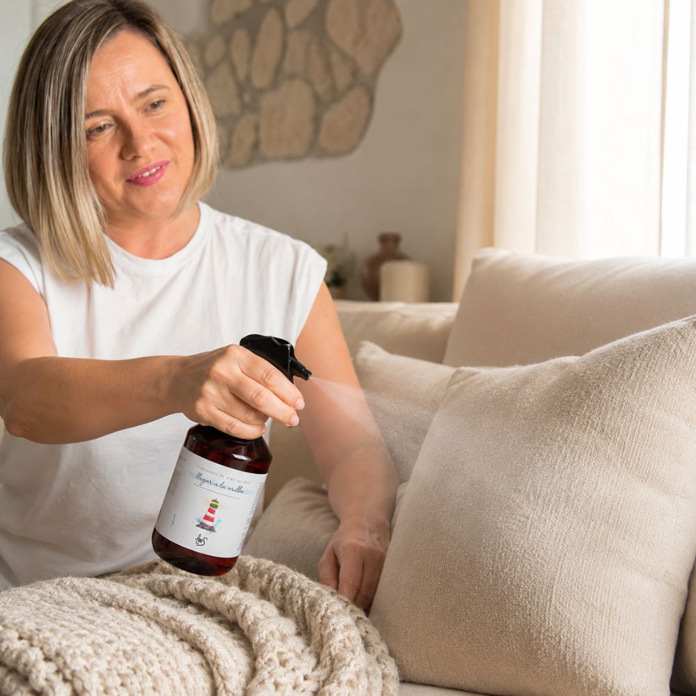 Ana Belén, fundadora de D'Arome, pulverizando spray textil eliminador de olor para tejidos Llegar a la Orilla de LoeS sobre sofá con manta de punto – D'Arome