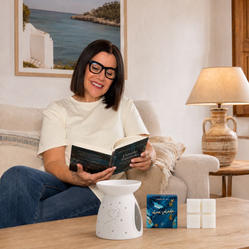 Mari Carmen, CEO y experta en aromas de D'Arome, disfruta de un momento de lectura junto a las tartaletas de cera Alma Salada de LoeS y su quemador en un salón con cuadro marino – D'Arome