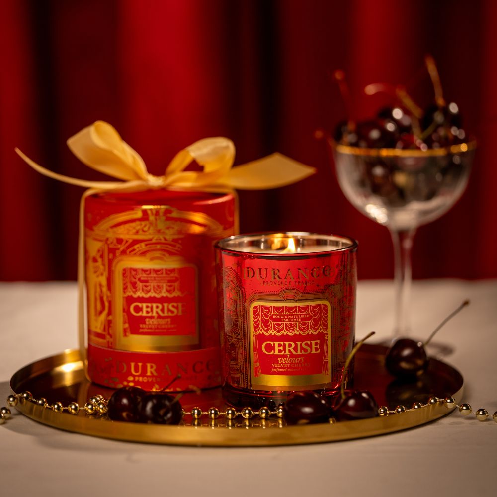Vela aromática cera natural Cereza Aterciopelada 300g de Durance encendida junto a su caja de regalo con lazo dorado y copa de cerezas frescas, sobre bandeja dorada – D'Arome