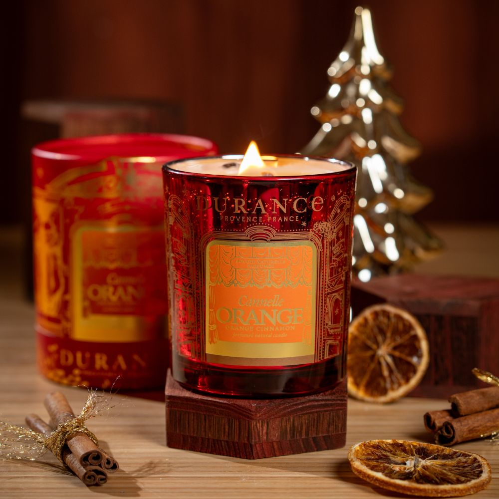 Vela aromática cera natural Naranja Canela 300g de Durance encendida sobre base de madera, con rodajas de naranja deshidratada y ramas de canela – D'Arome