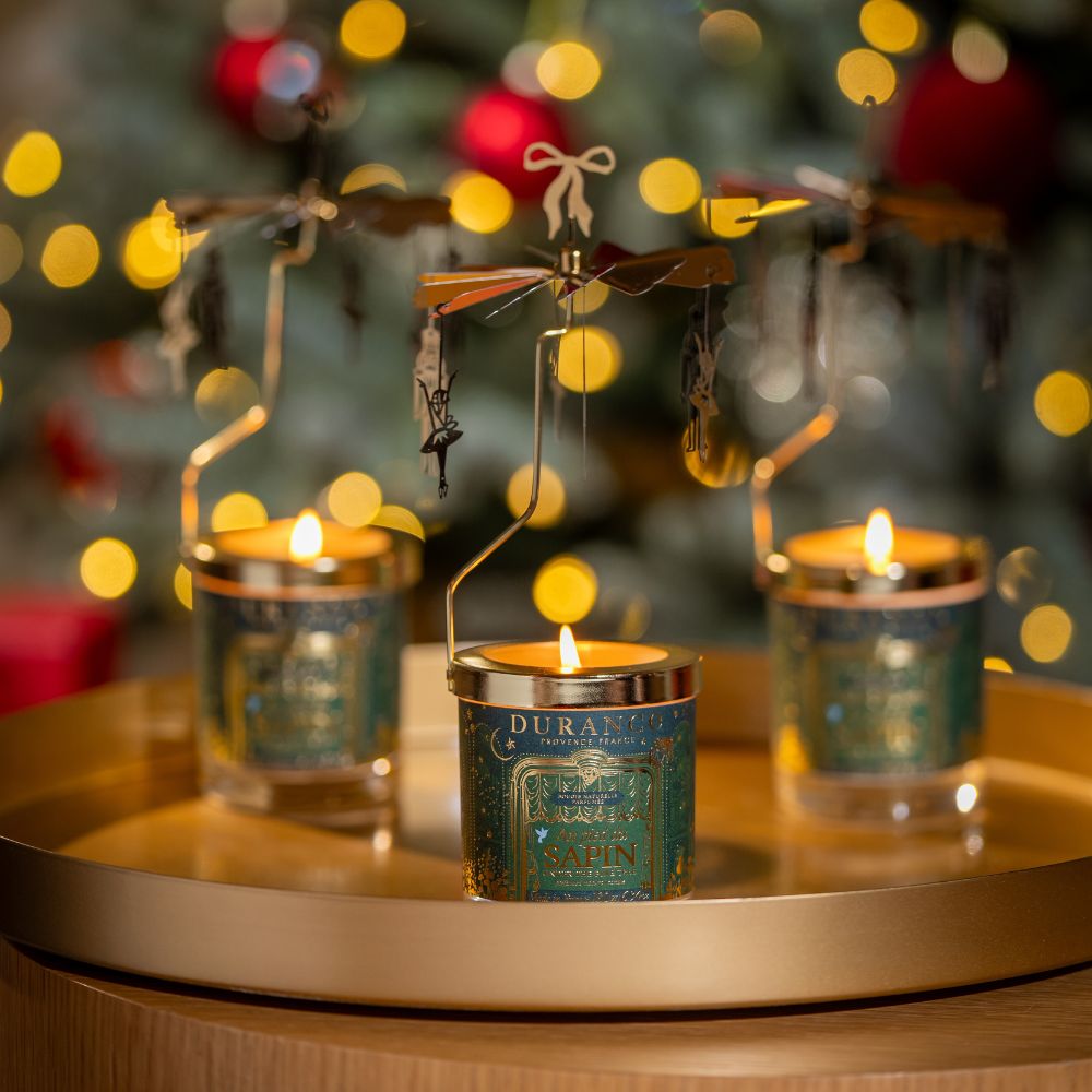 Set de tres velas carrusel Al Pie del Abeto 180gr de Durance encendidas sobre bandeja dorada, con figuras giratorias doradas y árbol de Navidad iluminado de fondo – D'Arome