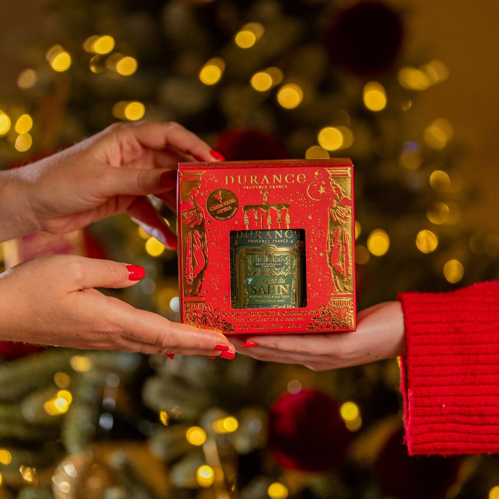 Manos con manicura roja sujetando la caja regalo de la vela carrusel Al Pie del Abeto 180gr de Durance frente a árbol de Navidad decorado – D'Arome
