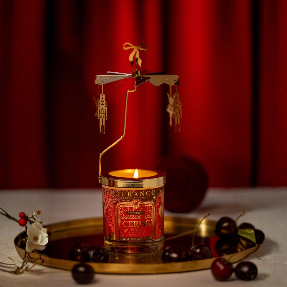 Vela carrusel Cereza Aterciopelada 180gr de Durance encendida sobre bandeja dorada con cerezas frescas y flor blanca, fondo de cortina roja navideña – D'Arome