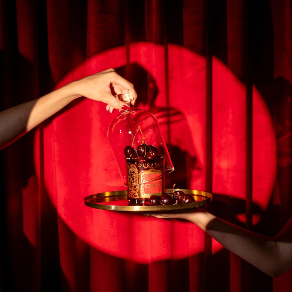 Mano cerrando una campana de cristal sobre la vela aromática Cereza Aterciopelada 300g de Durance y cerezas frescas, sobre bandeja dorada con foco de luz roja teatral – D'Arome