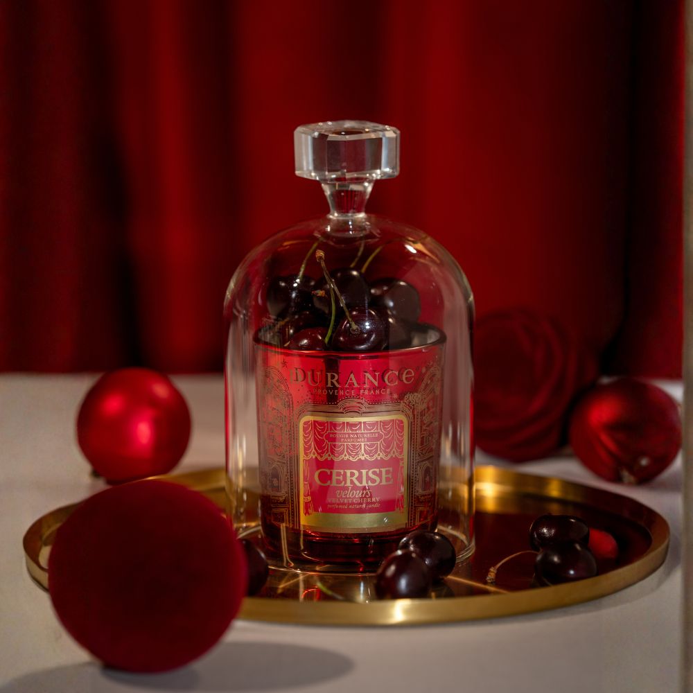 Primer plano de la vela aromática Cereza Aterciopelada 300g de Durance bajo campana de cristal con cerezas, rodeada de bolas de Navidad rojas de terciopelo – D'Arome