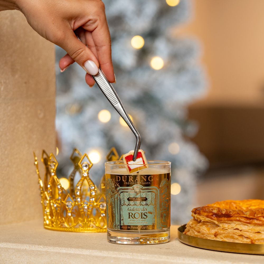 Mano extrayendo la haba sorpresa de la vela Pastel de Reyes 180gr de Durance con pinzas, junto a corona dorada y roscón de Reyes – D'Arome