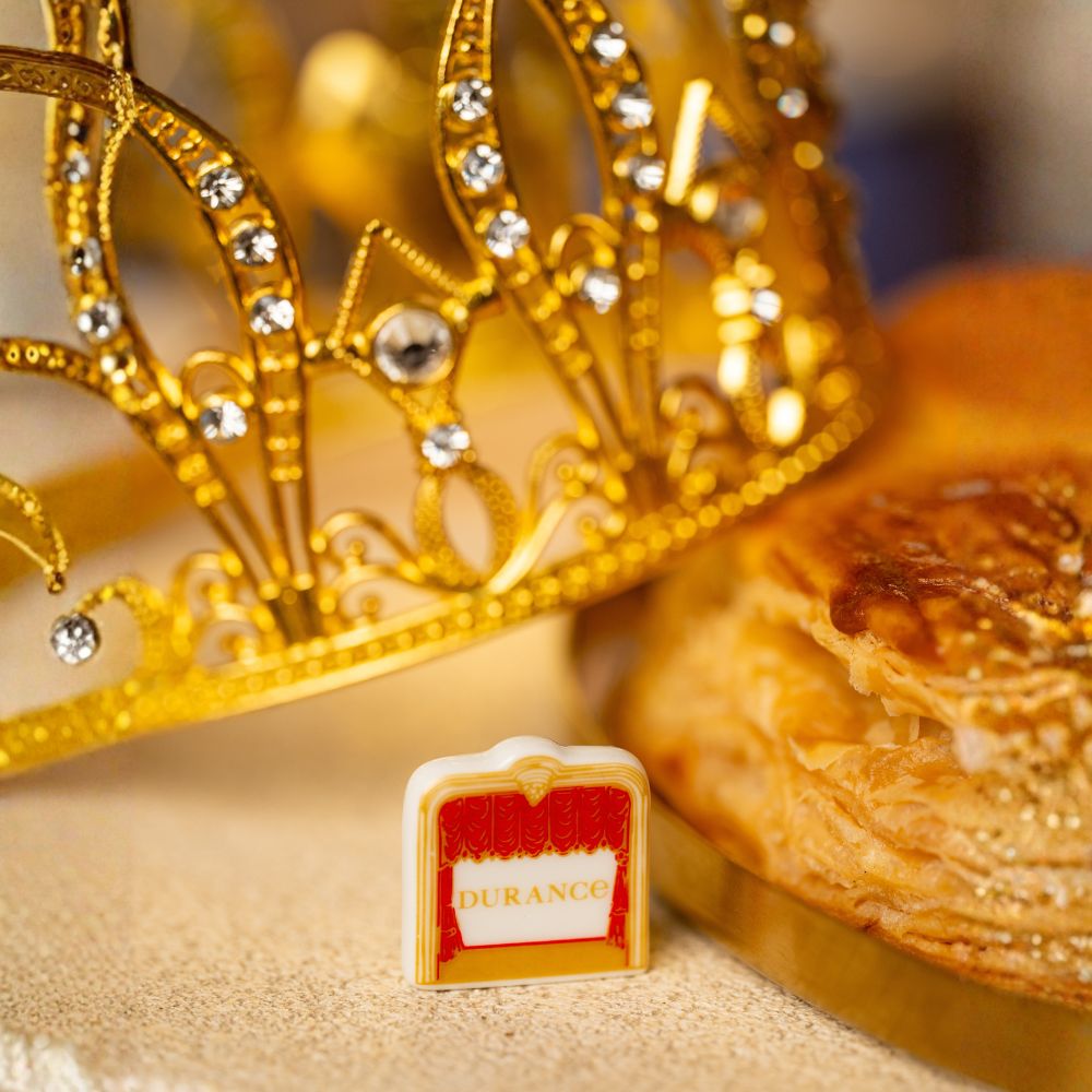 Detalle de la haba sorpresa de porcelana de la vela Pastel de Reyes de Durance, con diseño de cortina roja, junto a corona dorada y roscón de Reyes – D'Arome